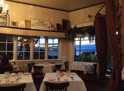 Cozy restaurant interior with white tablecloths, large windows, and a view of the ocean at sunset.