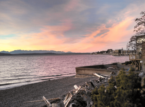 A serene beach scene at sunset, with mountains in the distance and colorful clouds reflecting on the water.