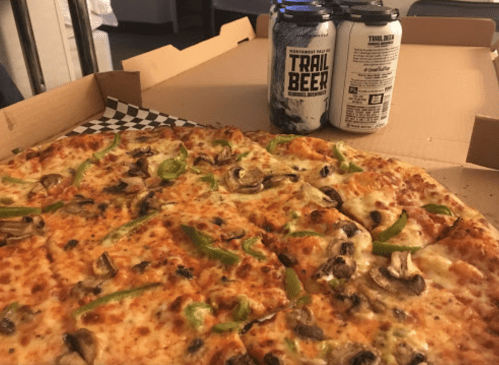 A pizza topped with green peppers and mushrooms, next to two cans of Trail Beer, in a pizza box.