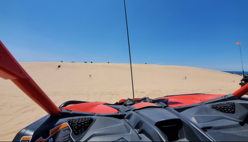 View from an ATV dashboard facing a sandy dune under a clear blue sky, with small figures in the distance.