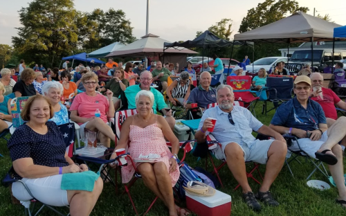 A group of smiling people sitting in lawn chairs at an outdoor event, enjoying drinks and socializing.