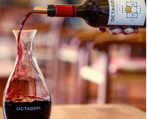 A hand pours red wine from a bottle into a glass decanter labeled "OCTAGON."