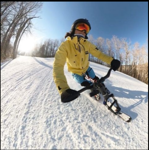 A person in a yellow jacket and helmet rides a snow bike on a snowy trail surrounded by trees.