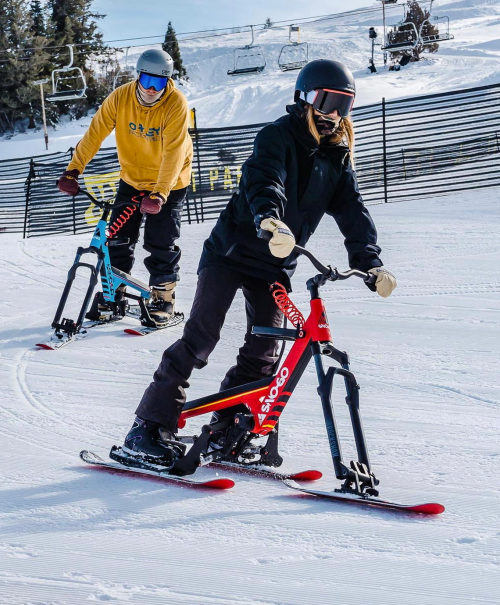 Two people riding snow bikes on a snowy slope, with ski lifts in the background and trees lining the area.