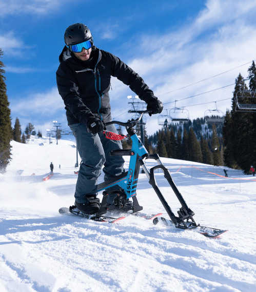 A person skiing on a snow bike, wearing a helmet and goggles, with a snowy mountain backdrop and ski lifts in the distance.