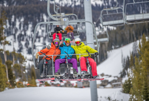 Three skiers in colorful outfits ride a ski lift, smiling against a snowy mountain backdrop.