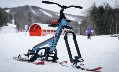 A ski bike with a blue frame and red skis stands on snow at a ski resort, with mountains in the background.