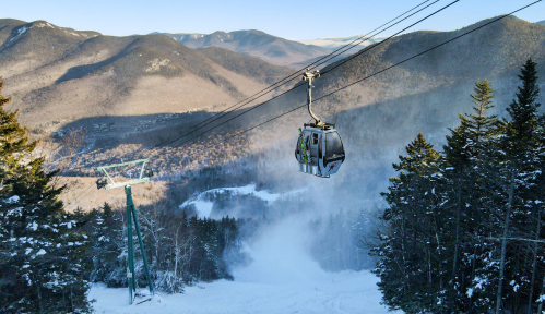 A ski gondola ascends a snowy mountain, surrounded by trees and distant peaks under a clear sky.