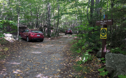 Two red cars parked near a trail sign in a wooded area with fallen leaves on the ground.