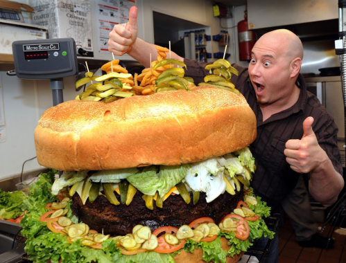 A man gives a thumbs up next to an enormous burger stacked with lettuce, pickles, and tomatoes in a kitchen setting.