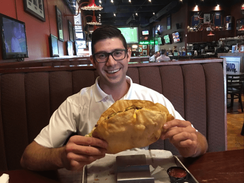A smiling man in a white shirt holds a large, stuffed taco in a restaurant, with TVs and patrons in the background.