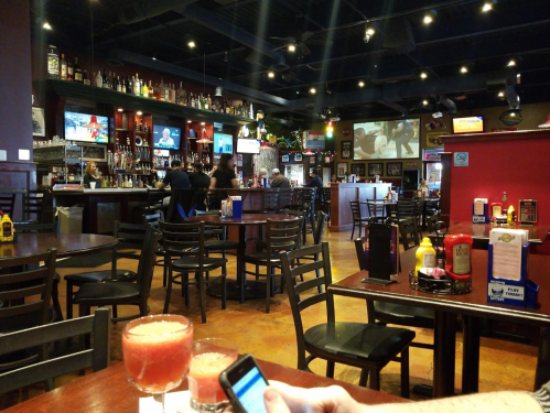 A lively bar scene with patrons, drinks on tables, and multiple TVs showing sports in a casual setting.