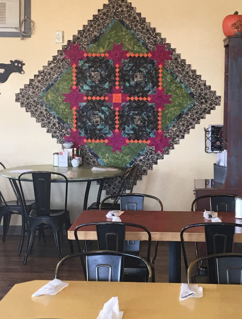 Colorful quilt hanging on a wall in a cozy restaurant, with tables and chairs set for dining.