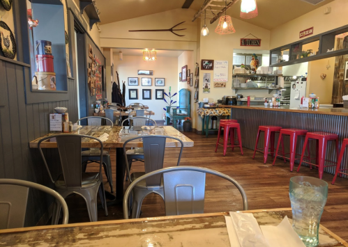 Cozy restaurant interior with wooden tables, metal chairs, and colorful decor, featuring a kitchen area in the background.