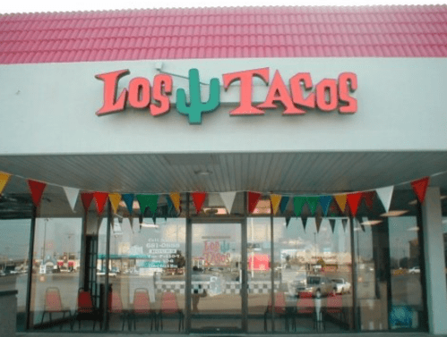 Exterior of "Los Tacos" restaurant with colorful flags and a red roof, featuring large signage.