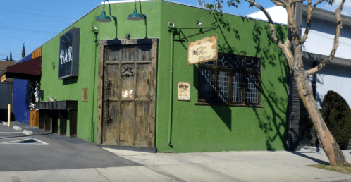 A green building with a wooden door and decorative signs, featuring a bar entrance and nearby trees.