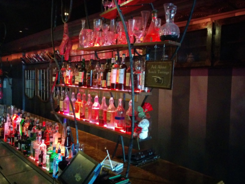 A dimly lit bar shelf displaying various bottles and glassware, illuminated with red lights.