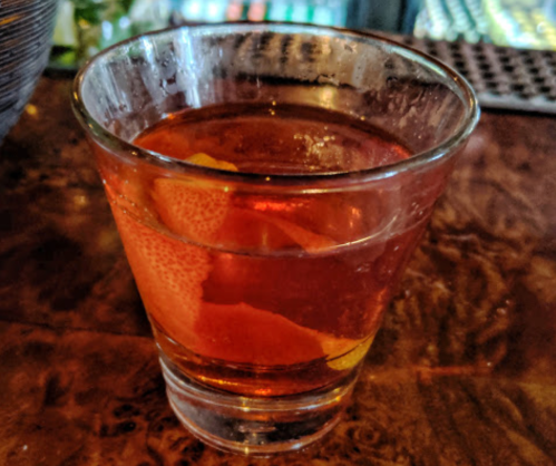 A glass of amber-colored cocktail with orange peel garnish, sitting on a wooden bar surface.
