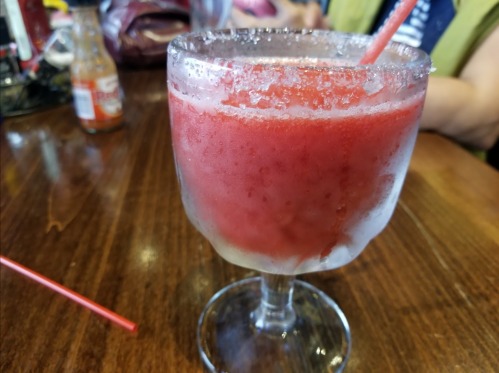 A frozen red drink in a frosted glass, with a straw, on a wooden table. Condiments are visible in the background.