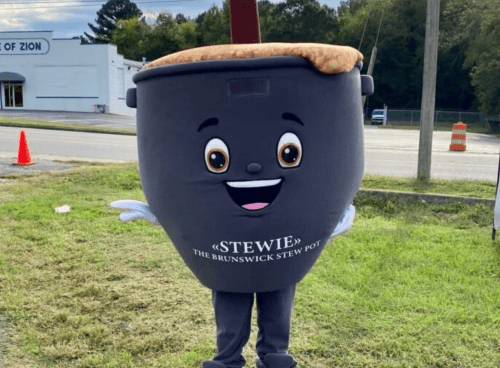 A cheerful mascot named Stewie, resembling a stew pot, stands on a grassy area with a smiling face and arms.