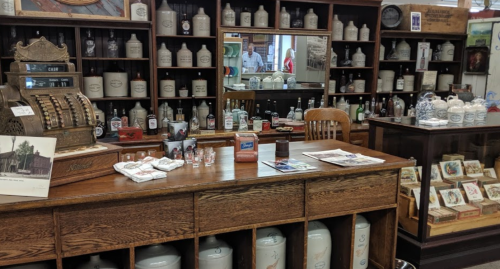 A vintage store display featuring glass bottles, an antique cash register, and various historical items on wooden shelves.