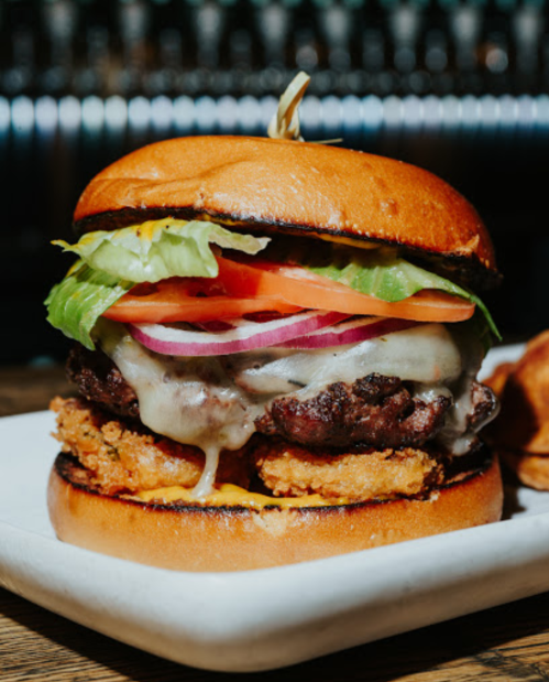A close-up of a juicy burger with lettuce, tomato, onion, cheese, and crispy elements on a toasted bun.