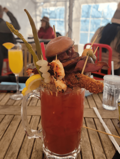 A tall glass of Bloody Mary garnished with shrimp, bacon, a mini burger, asparagus, and lemon, set on a wooden table.