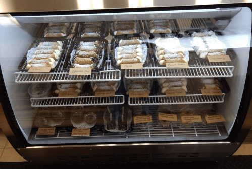 A display case filled with various desserts, including cannoli and donuts, each labeled with their flavors.