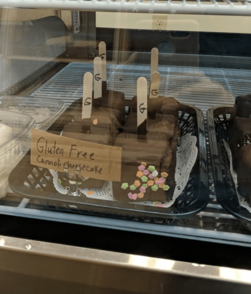 Gluten-free cannoli cheesecake slices displayed in a glass case, topped with colorful sprinkles and labeled.