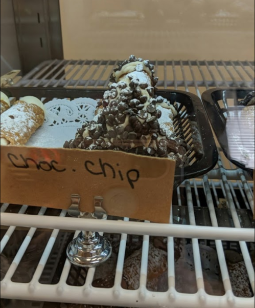 A display case with a chocolate chip dessert topped with powdered sugar and chocolate chips, alongside other pastries.