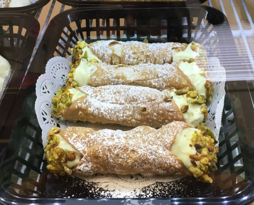 Three cannoli filled with cream and topped with crushed pistachios, dusted with powdered sugar, displayed in a basket.