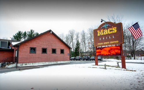 A wooden building with a sign reading "Mac's Grill" and an American flag, surrounded by snow and trees.