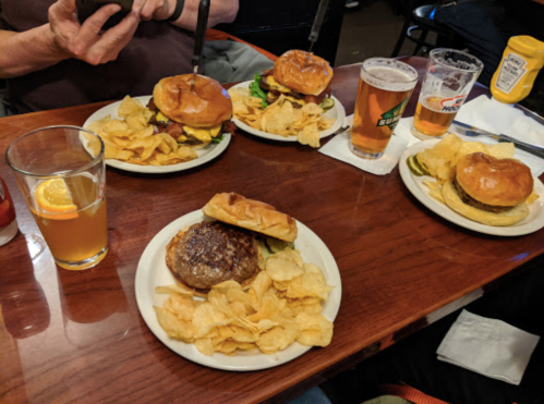A table with four burgers, plates of chips, two glasses of beer, and a drink with an orange slice.