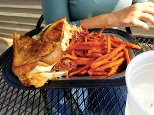 A plate with a grilled sandwich and sweet potato fries, set on a table with a person’s hand in the background.