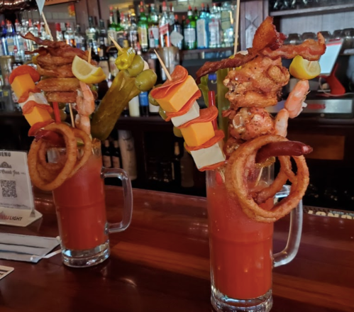 Two large cocktails with elaborate garnishes, including shrimp, bacon, cheese, and vegetables, on a bar counter.