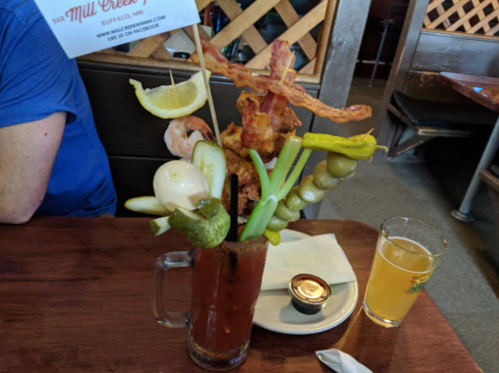 A tall Bloody Mary garnished with bacon, shrimp, pickles, olives, and celery, alongside a glass of beer.