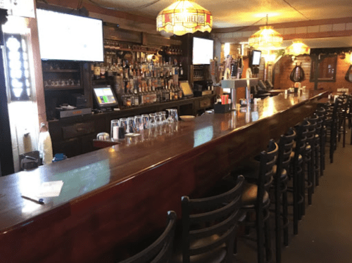 A wooden bar with empty stools, shelves of liquor, and screens in a cozy pub setting.
