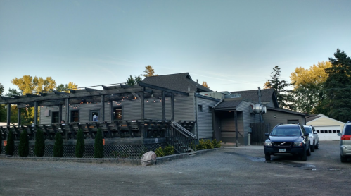 A rustic building with a wooden deck and outdoor seating, surrounded by trees and parked cars.