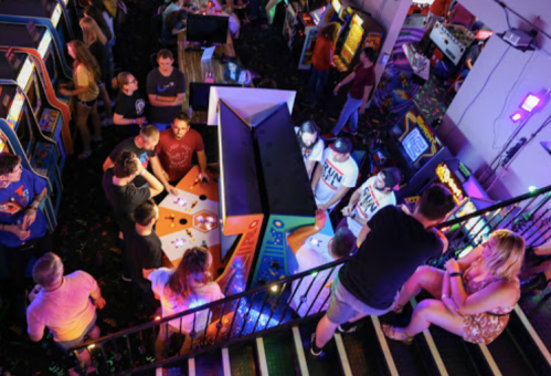A vibrant arcade scene with people playing games, colorful lights, and a staircase in the foreground.
