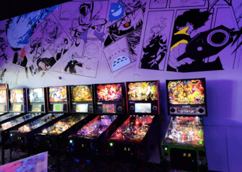 A vibrant arcade with colorful pinball machines and a mural featuring anime characters on the wall.