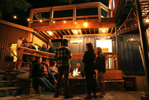 A lively outdoor gathering at night, with people socializing around a fire pit and string lights illuminating the space.