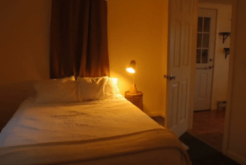 Cozy bedroom with a bed, warm lighting, and a door leading to another room. Soft colors create a relaxing atmosphere.