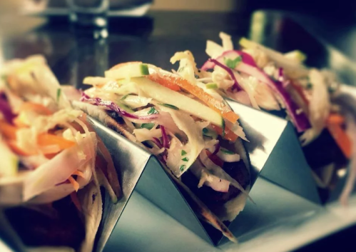 Three tacos in a metal holder, topped with colorful slaw and fresh vegetables.
