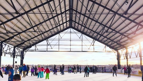 A lively outdoor ice skating rink with people of all ages skating under a large, open-roof structure at sunset.