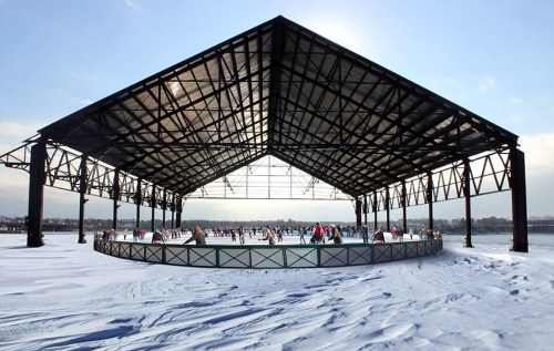 A large open-air ice skating rink under a metal roof, surrounded by snow and skaters enjoying the winter day.