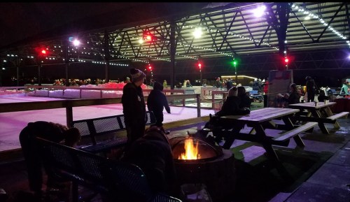 A cozy outdoor ice skating rink at night, with colorful lights and people enjoying the atmosphere around a fire pit.