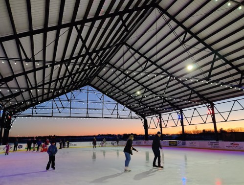 People skating on an ice rink under a large, open structure with a sunset in the background.