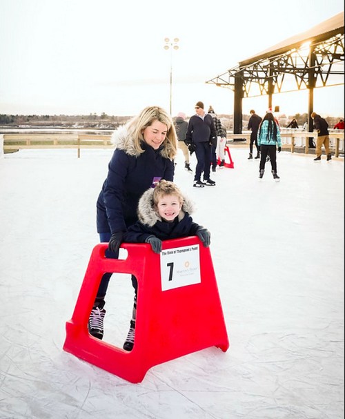 A woman helps a child skate on ice using a red skating aid, with other skaters in the background and a sunset.