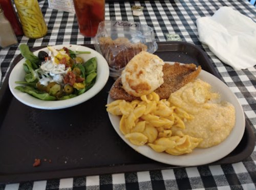A tray with a salad, fried fish, macaroni and cheese, grits, and a biscuit on a checkered tablecloth.