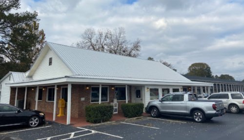 A one-story building with a metal roof, surrounded by parked cars and trees under a cloudy sky.